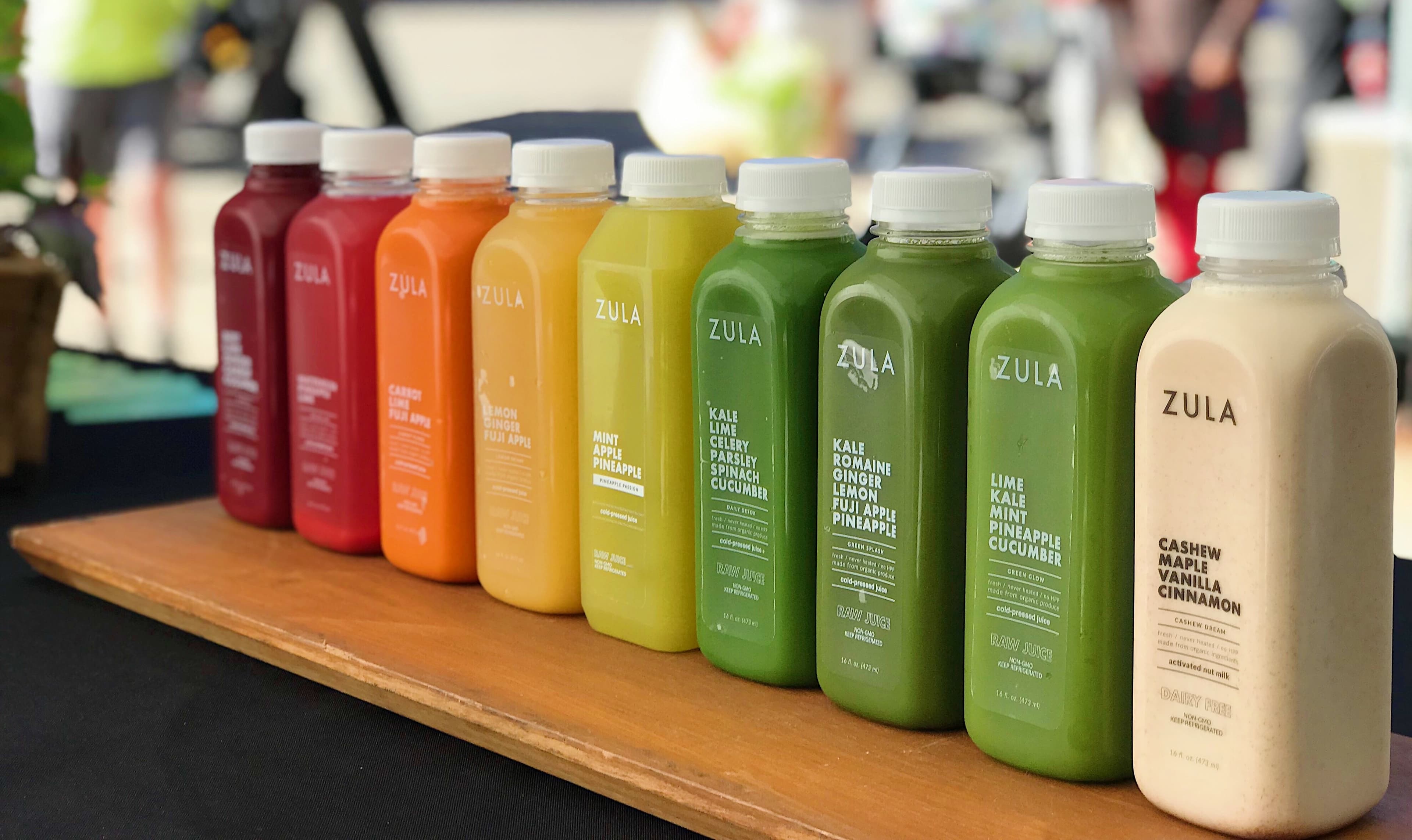 Historical Zula Juice bottles in a range of colors on a wooden counter beside fresh produce.